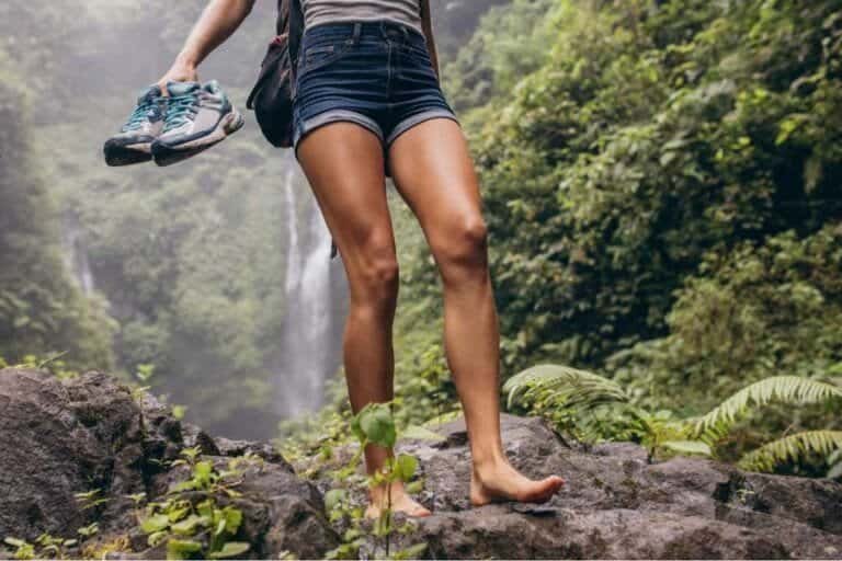 A woman standing barefoot on rocks near a waterfall in the jungle.