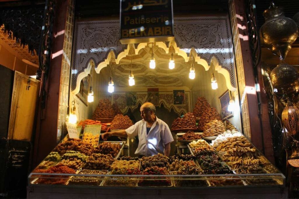 Backpacking trip to Marrakesh visiting the souks. A man in a market stall serving sweets and deserts with golden lights above him