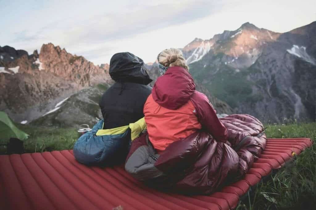Backpackers sitting on a camping mat in front of mountain landscape, enjoying outdoor adventure and exploring nature.
