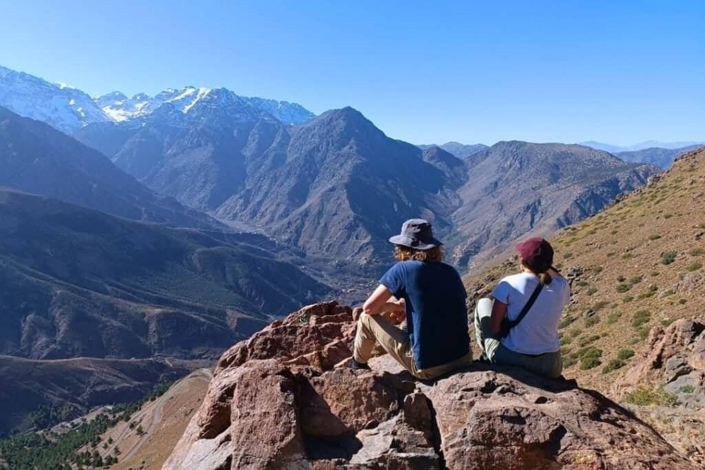 A breathtaking mountain landscape with two backpackers sitting on a rock, enjoying the view of rugged peaks and deep valleys, emphasizing adventure, hiking, and travel experiences.