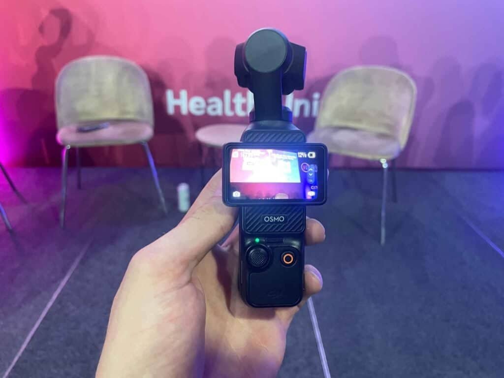 Cinematic shot of a gimbal stabilizer capturing a conference or seminar setting with empty chairs against a pink wall with "Health &" visible in the background.