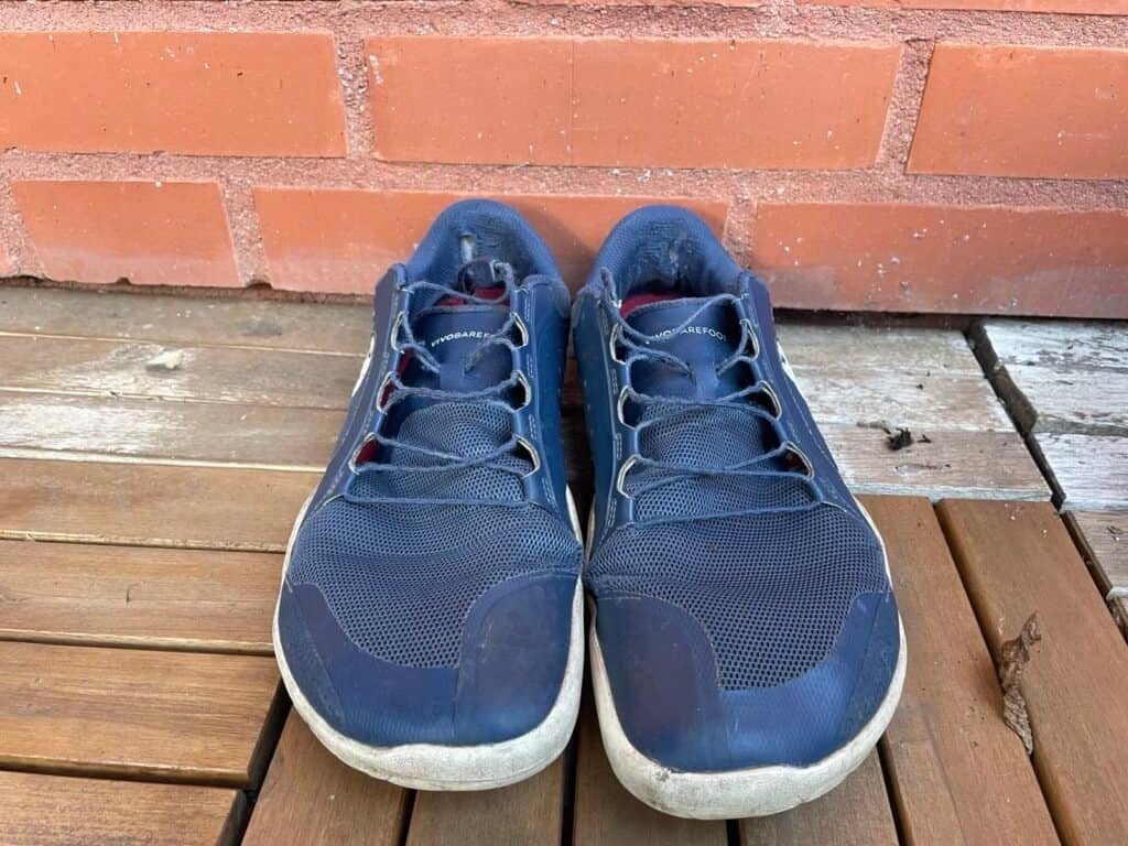 Vivobarefoot running shoes Comfortable blue trail running shoes placed on a wooden deck against a red brick wall, ideal for backpackers and outdoor adventures.