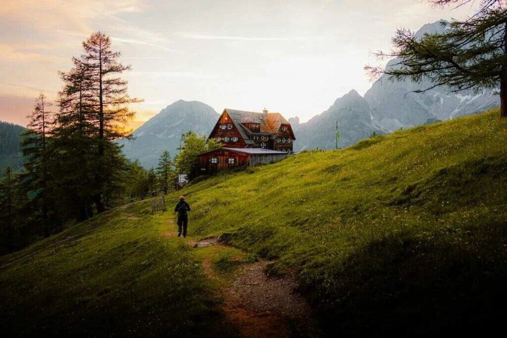 Serene mountain landscape with cozy wooden cabin, lush green grass, tall trees, and majestic peaks at sunset, perfect for outdoor adventures and backpacking trips with Barefoot Backpackers.