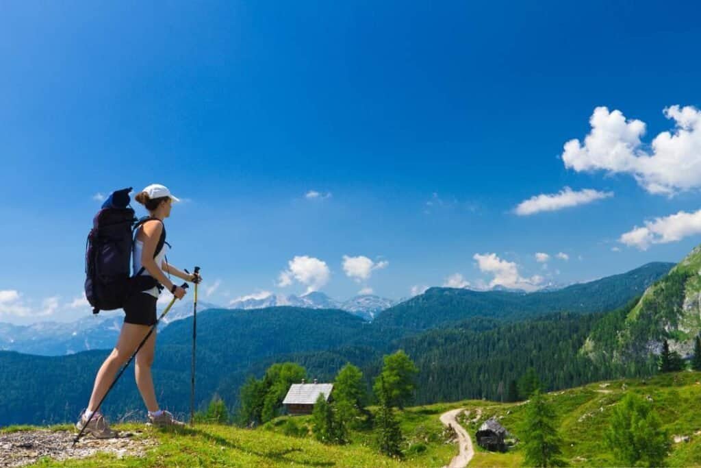 Bright female backpacker hiking on mountain trail with scenic landscape, blue skies, and lush greenery, experiencing outdoor adventure and nature travel in the mountains.