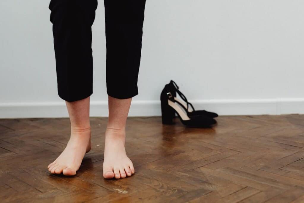 Barefoot person standing on wooden floor with black heels beside, eco-conscious lifestyle, mindful travel, barefoot exploration. L