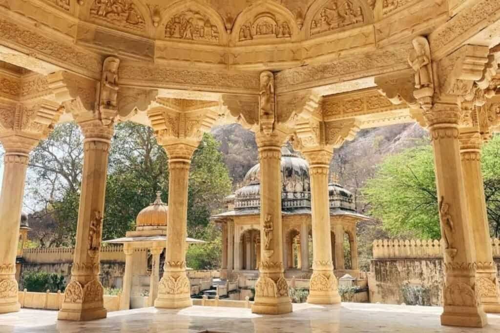 Jaipur-Desktop Intricate stone carvings and architectural details of an Indian temple with carved pillars and domed structures, set against lush greenery and mountains in the background.