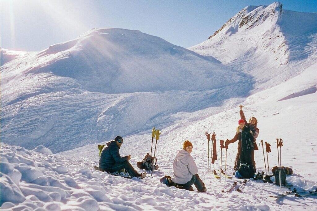 Cold snow-covered mountain landscape with three hikers resting and preparing gear in bright winter sunlight.