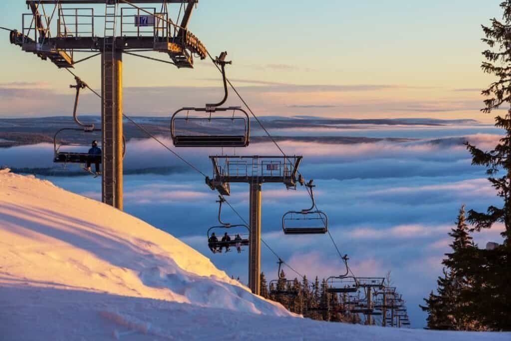 Gondola ski lift ascending above snow-covered slopes during sunset with clouds below.