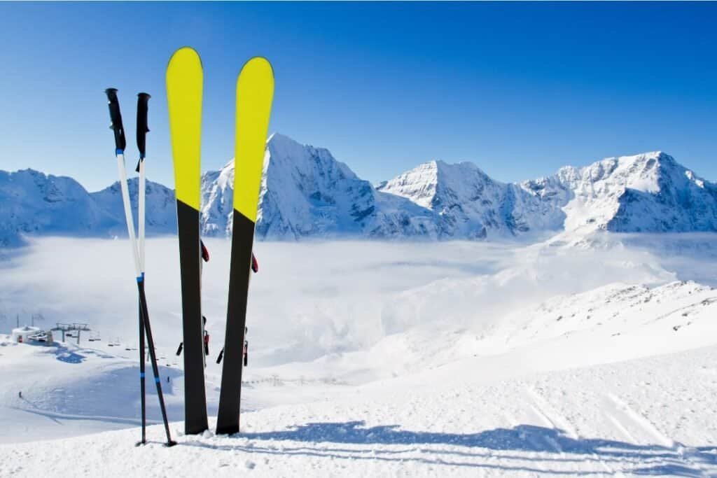 Vibrant yellow skis and poles standing upright in snow with majestic snow-capped mountains in the background under a clear blue sky.