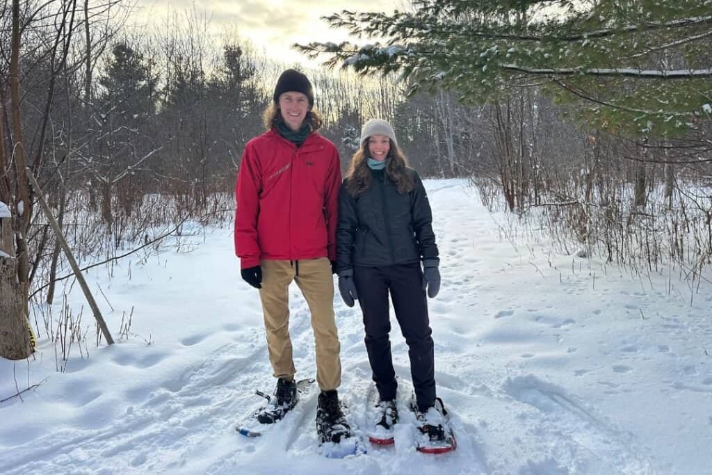 Snowshoeing Two smiling travelers in snowshoes enjoy a peaceful winter hike through a snowy forest landscape. They are dressed warmly for the cold, surrounded by bare trees and evergreen branches, embodying mindful exploration of nature's winter beauty. This image inspires sustainable outdoor adventures in harmony with the environment.