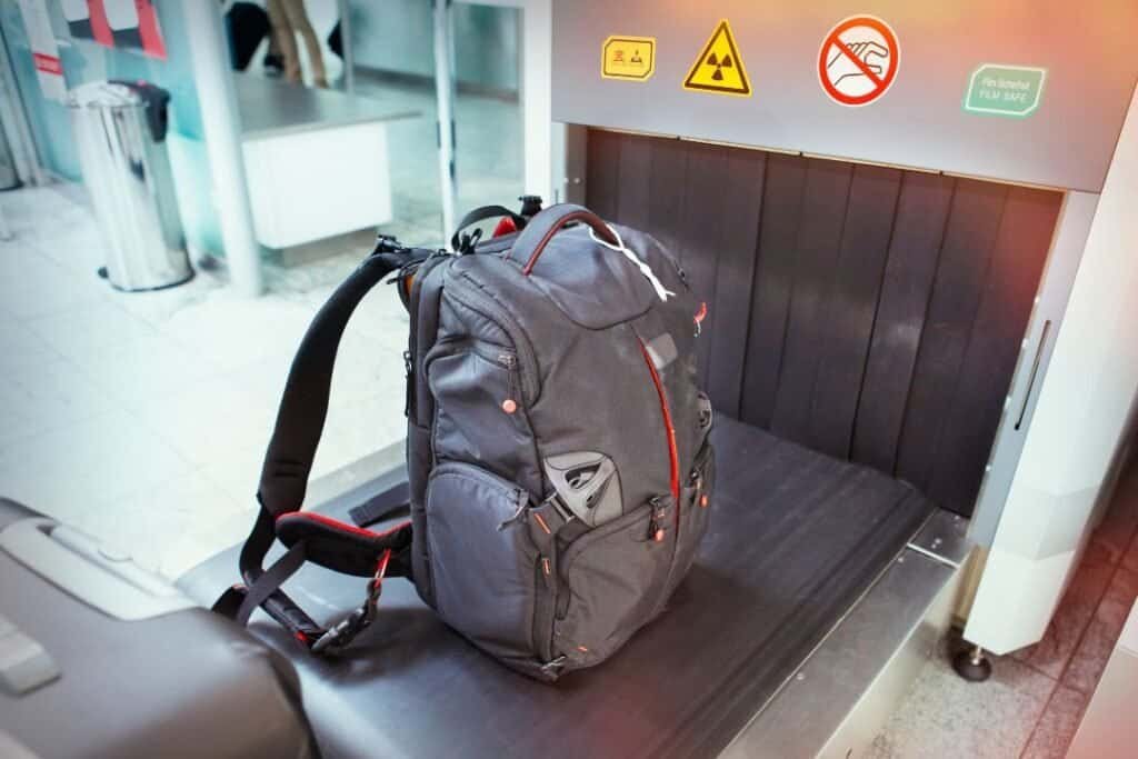 Travel Gear Bag. A black backpack on a baggage scanner conveyor at airport security, with safety warning signs above, emphasizing mindful and sustainable travel preparedness.