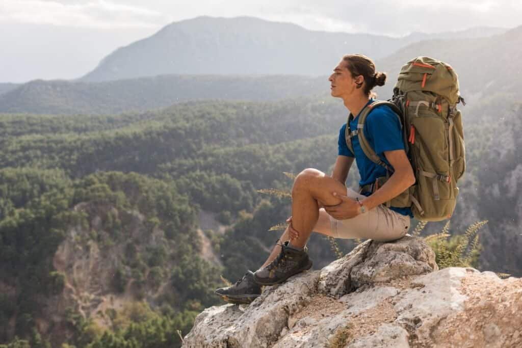 young man sitting peacefully on a rock overlooking lush green mountains, with a backpack, embracing mindful adventure and nature connection.