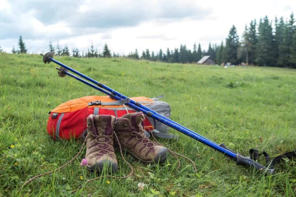 Backpacking gear including hiking boots, trekking poles, and a backpack on a grassy field with forest in the background.
