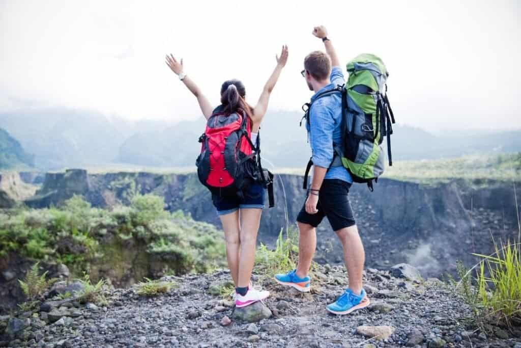 Vibrant hikers with 50L backpack celebrating on a mountain trail with lush green landscape and volcanic crater in the background.