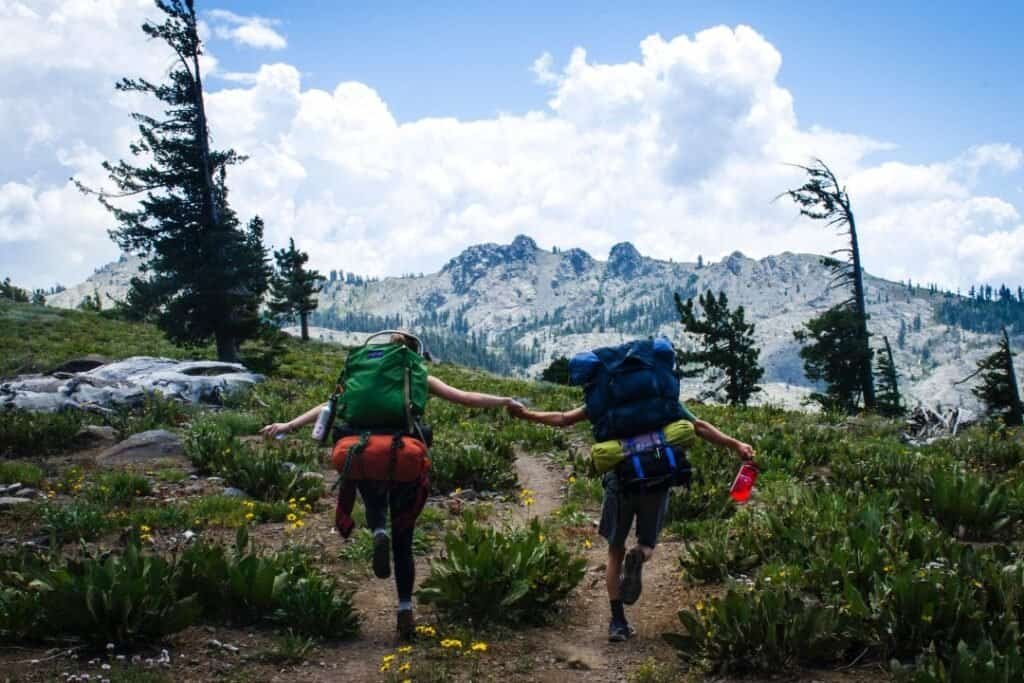 Young hikers holding hands on a mountain trail with backpacks, surrounded by lush greenery, rocky peaks, and a bright blue sky.