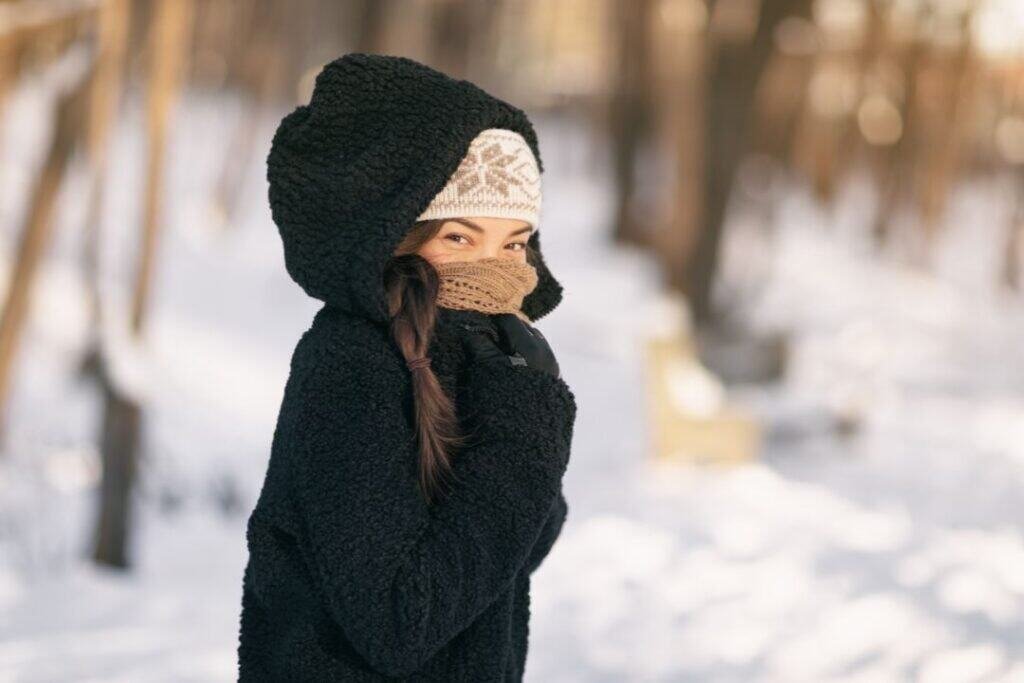 Cozy winter woman bundled in warm hat, scarf, and jacket in snowy forest setting, embracing mindful and sustainable winter travel adventures.