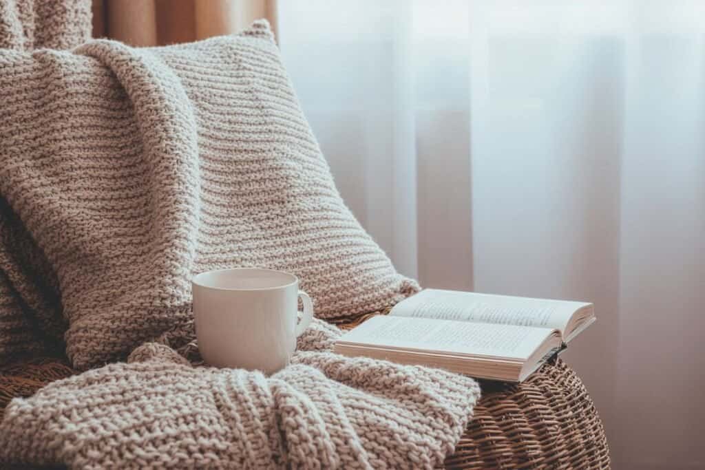 Cozy reading nook with knitted 100% wool throw blanket, cup of tea, and open book near a sunlit window for mindful, restful moments.