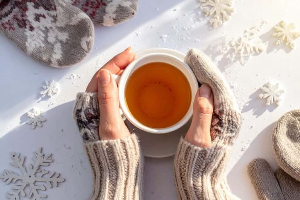 Comforting winter tea in cozy 100% Wool gloves on snow-covered surface with snowflake decorations, evoking warmth and mindful relaxation.