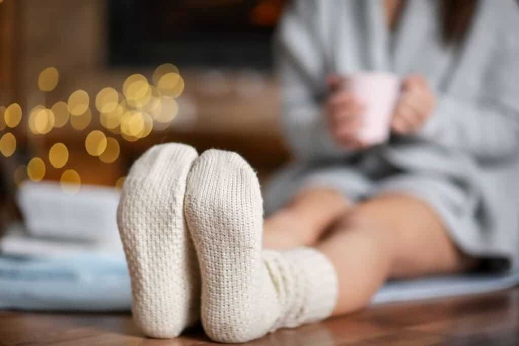 Cozy feet in warm knitted high quality socks and a person relaxing with a cup in a cozy, dimly lit home setting.