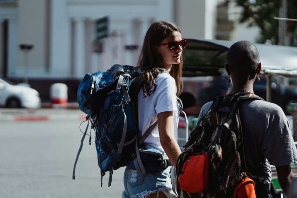 Young woman with a backpack and sunglasses talking to a person with a backpack on a city street, embracing mindful travel and adventures.