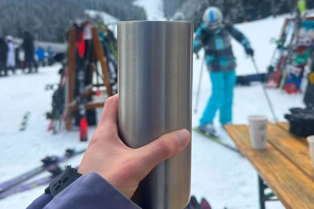 A stainless steel reusable travel mug held by a person at a snowy ski resort, with skiers, skis, and a mountain backdrop in the background.