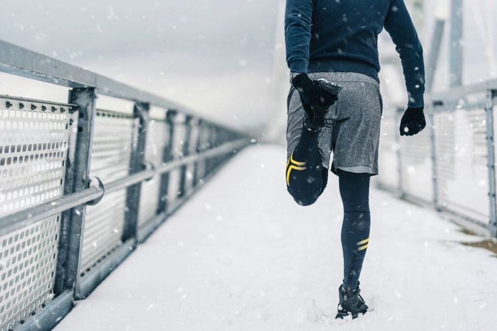 Dressed for winter running on a snow-covered bridge, in a cozy hoodie, shorts layered over leggings, and gloves, embracing outdoor adventure in cold weather using compression socks for men.