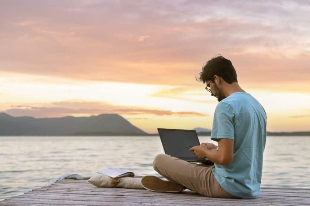 Young man working on a laptop by the water at sunset, embracing mindful remote work with digital nomad essentials.