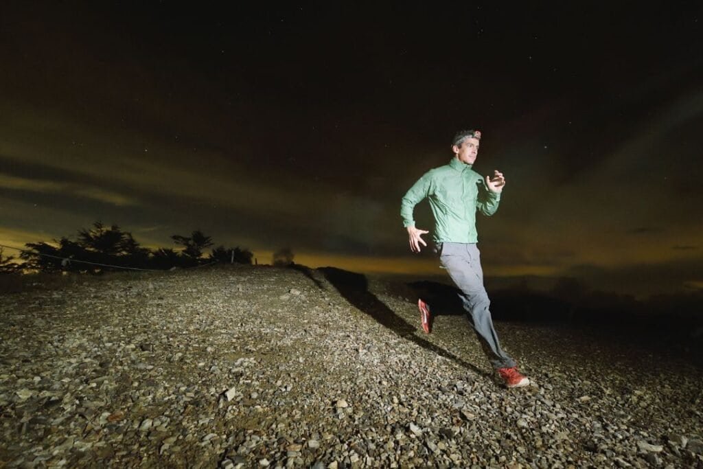 A person trail running at night on a rocky hill under a starry sky with northern lights.