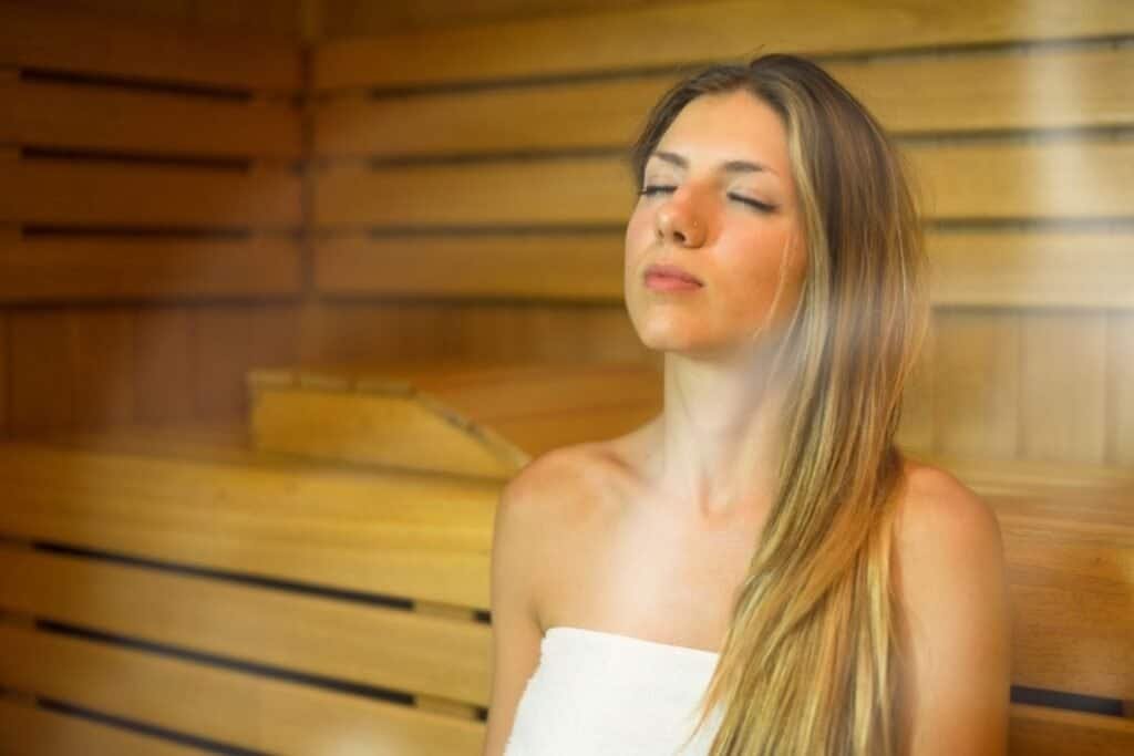 Calm woman enjoying a spa sauna in a wooden room with her eyes closed and steam around her, promoting mindfulness and relaxation.