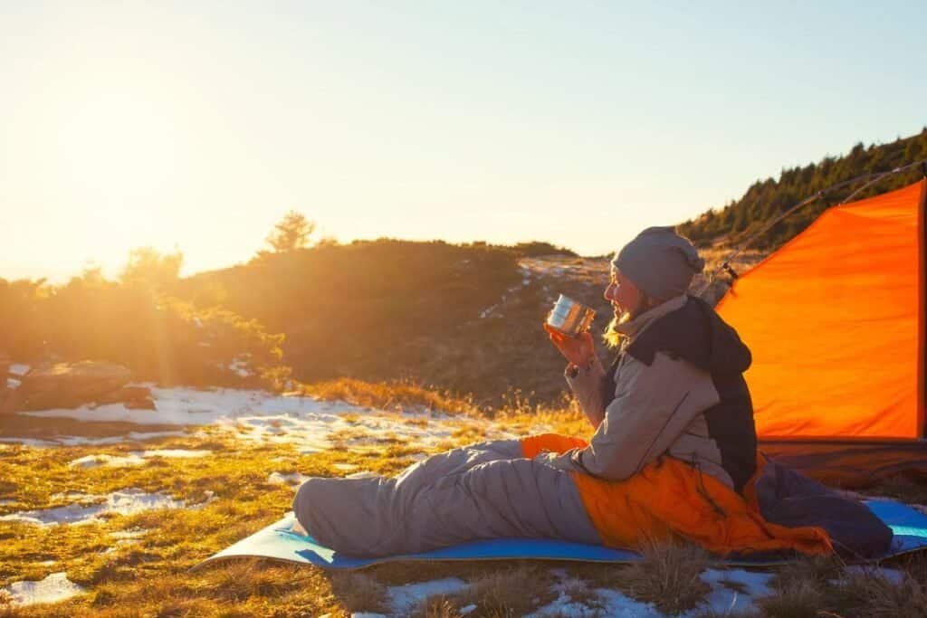 A person relaxing outdoors with a silk sleeping bag liner near a tent during sunset, emphasizing sustainable travel and conne.