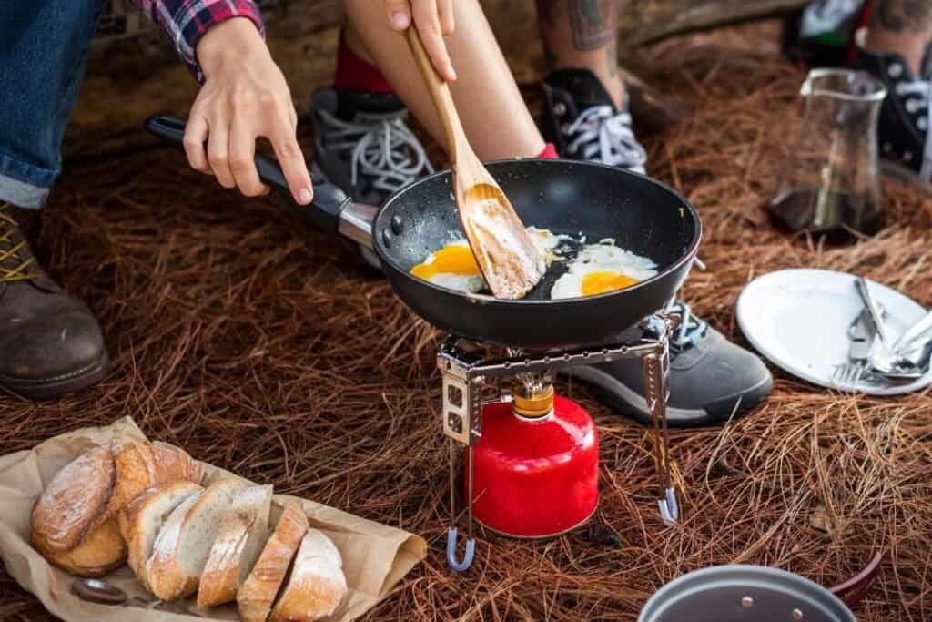 Crouching on a stool using a portable camping stove in use during a mindful outdoor breakfast, emphasizing connection with nature and sustainable travel.