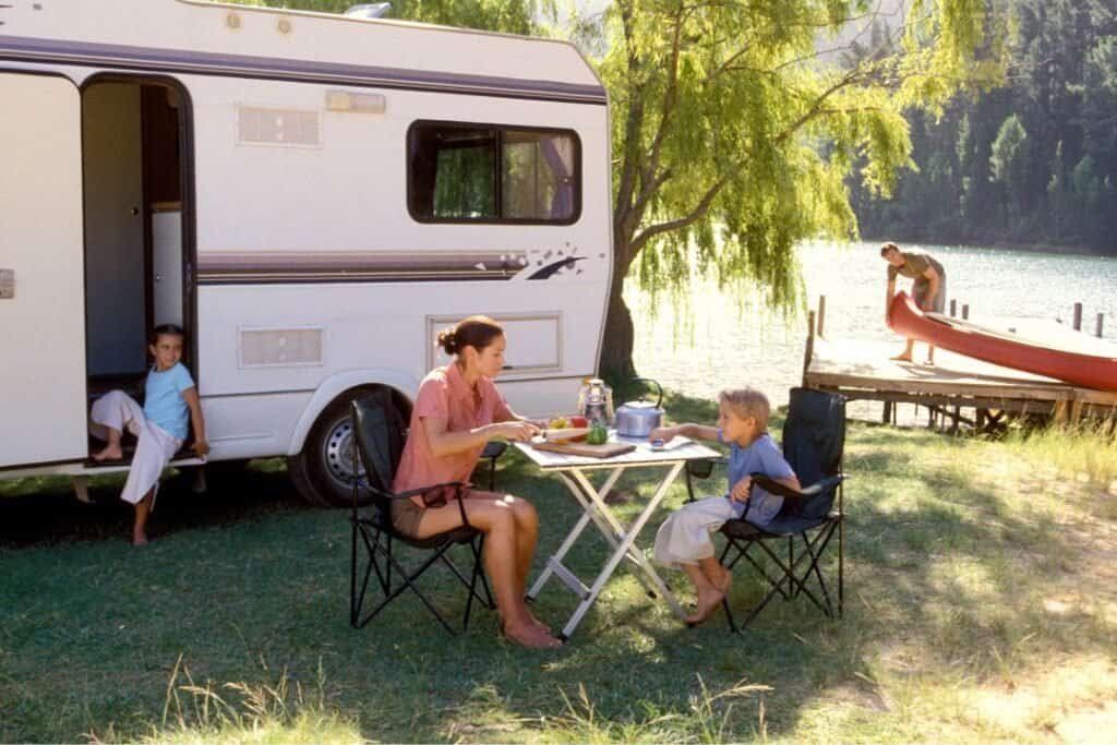 A family relaxing at a campsite near a tranquil lake, with a camper trailer, a table for outdoor dining, and a canoe on the s.