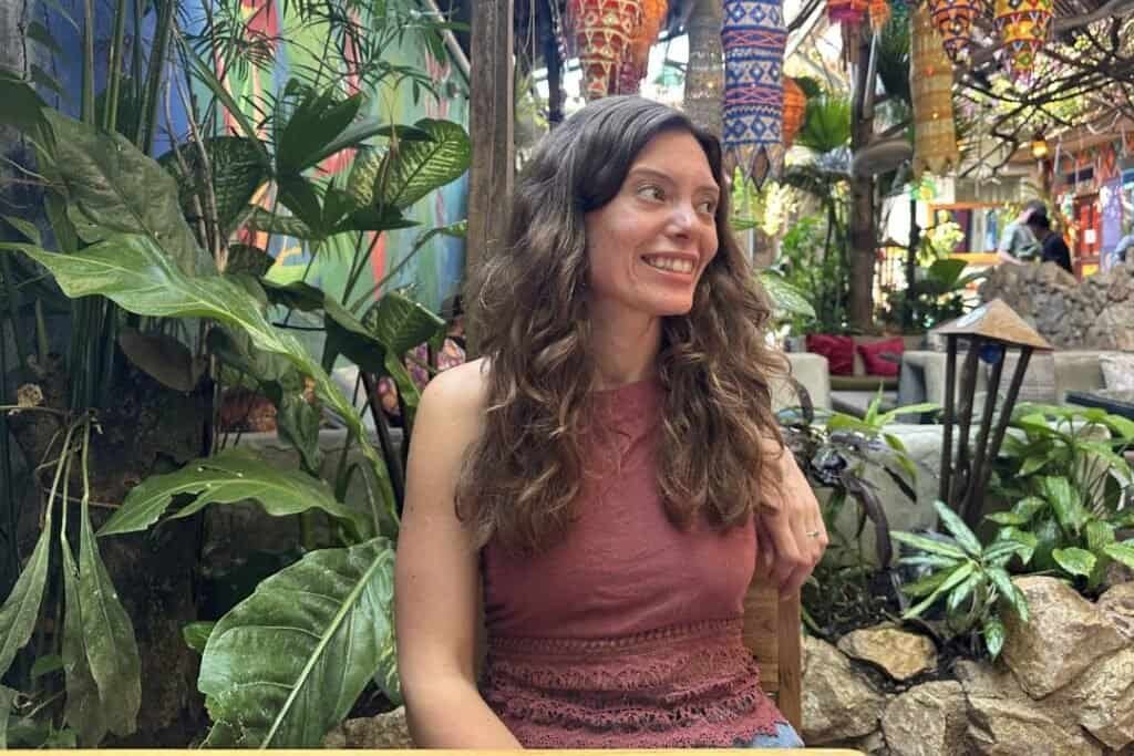 A woman with long, wavy hair smiling in a lush, tropical garden filled with vibrant plants and colorful decorations.